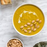A bowl of creamy pumpkin soup, half of a pumpkin cabotia, roasted pumpkin seeds and rosemary bread