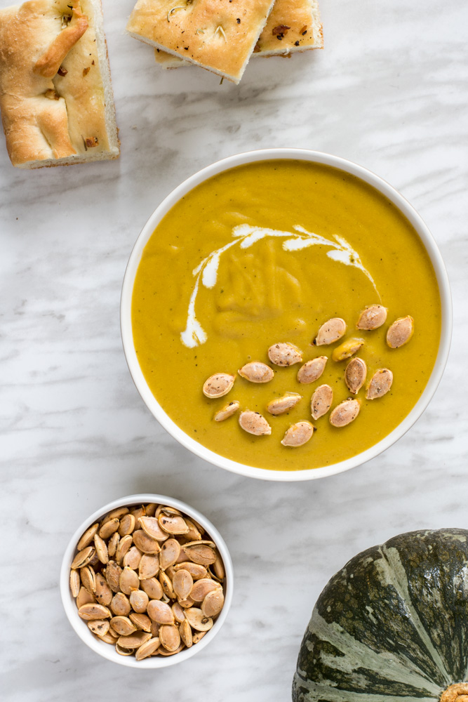 A bowl of creamy pumpkin soup, half of a pumpkin cabotia, roasted pumpkin seeds and rosemary bread