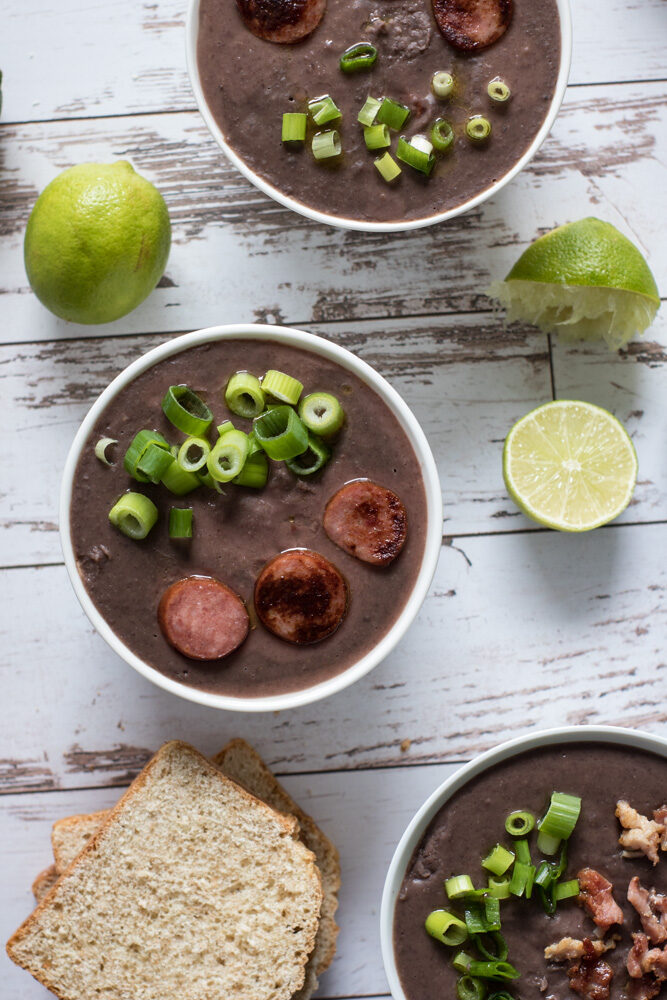 Bowls of bacon black bean soup with bread, lime, green onions, sausage and bacon.