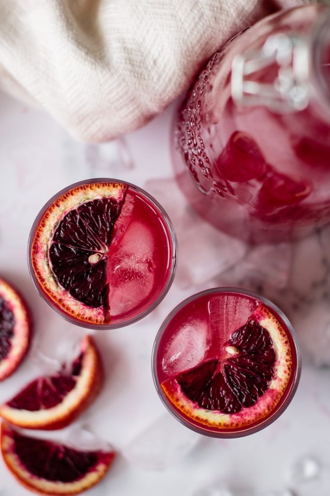 blood orange lemonade in two glasses and jar, with blood oranges on the top of the glass, ice, and slices of orange on the counter