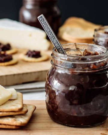 a jar with red onion chutney with a spoon inside, with crackers and cheese on the side, over a wooden board.