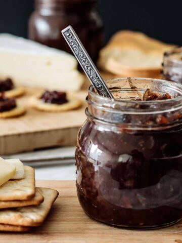 a jar with red onion chutney with a spoon inside, with crackers and cheese on the side, over a wooden board.