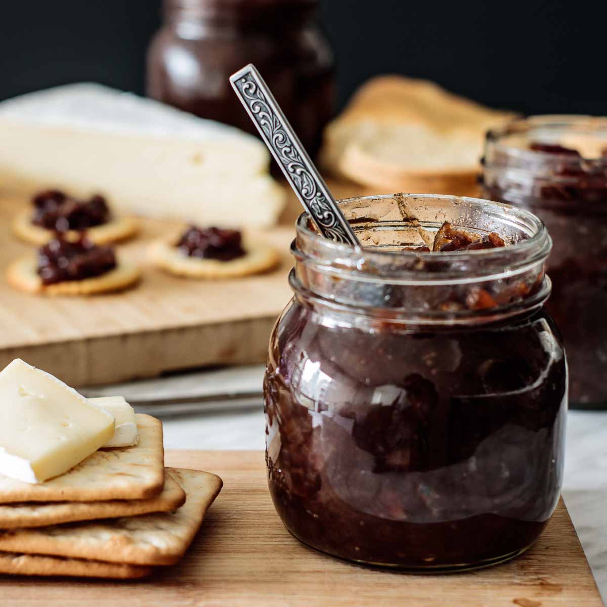 A jar full of onion chutney with a spoon inside, and crackers with cheese on its side, over a wooden board.