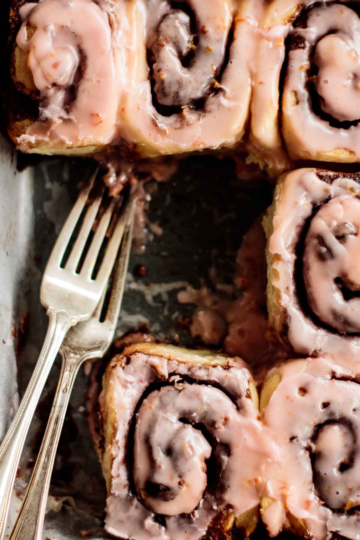 Blood orange cinnamon rolls with pink glaze and two forks on a baking pan.