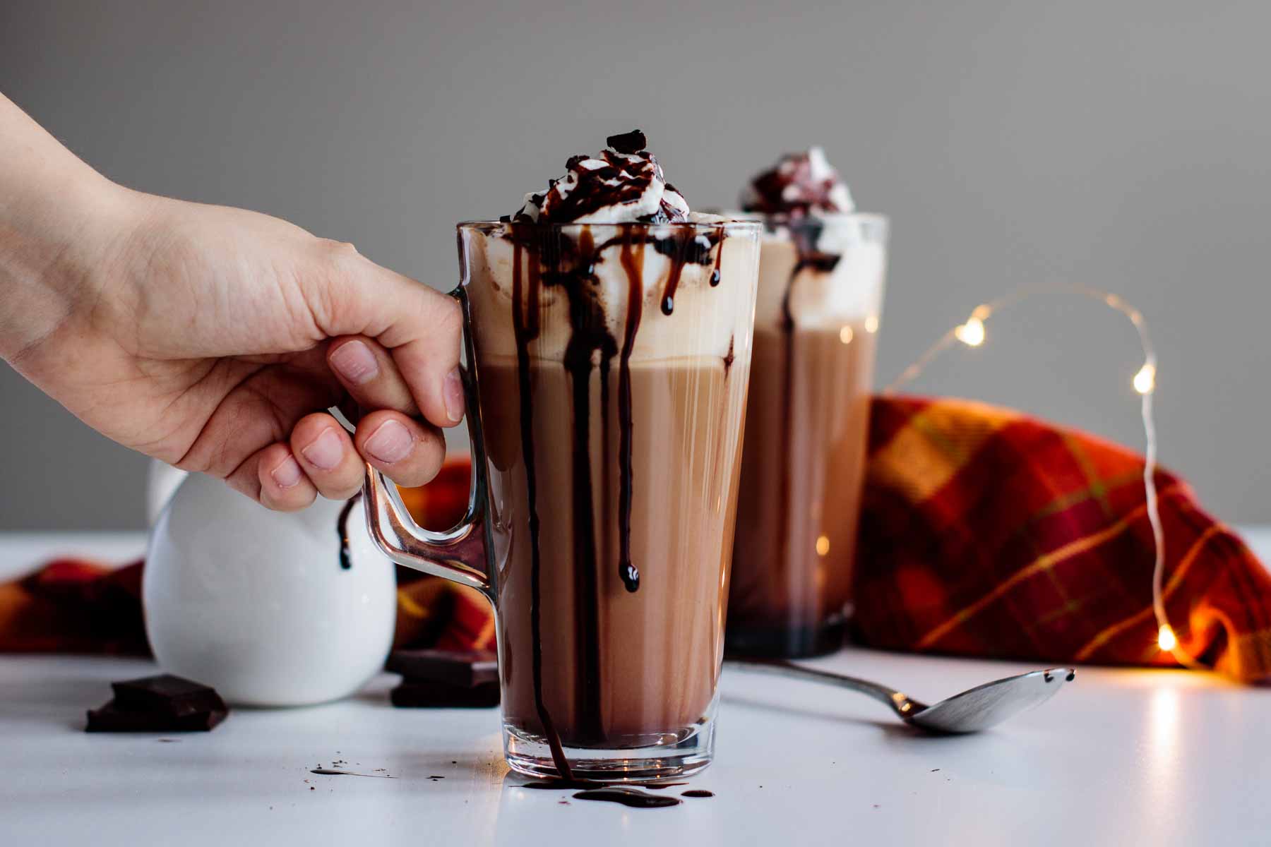 drinks ready, over a white table, with mocha sauce and lights in the background, and a hand ready to grab the closer glass cup