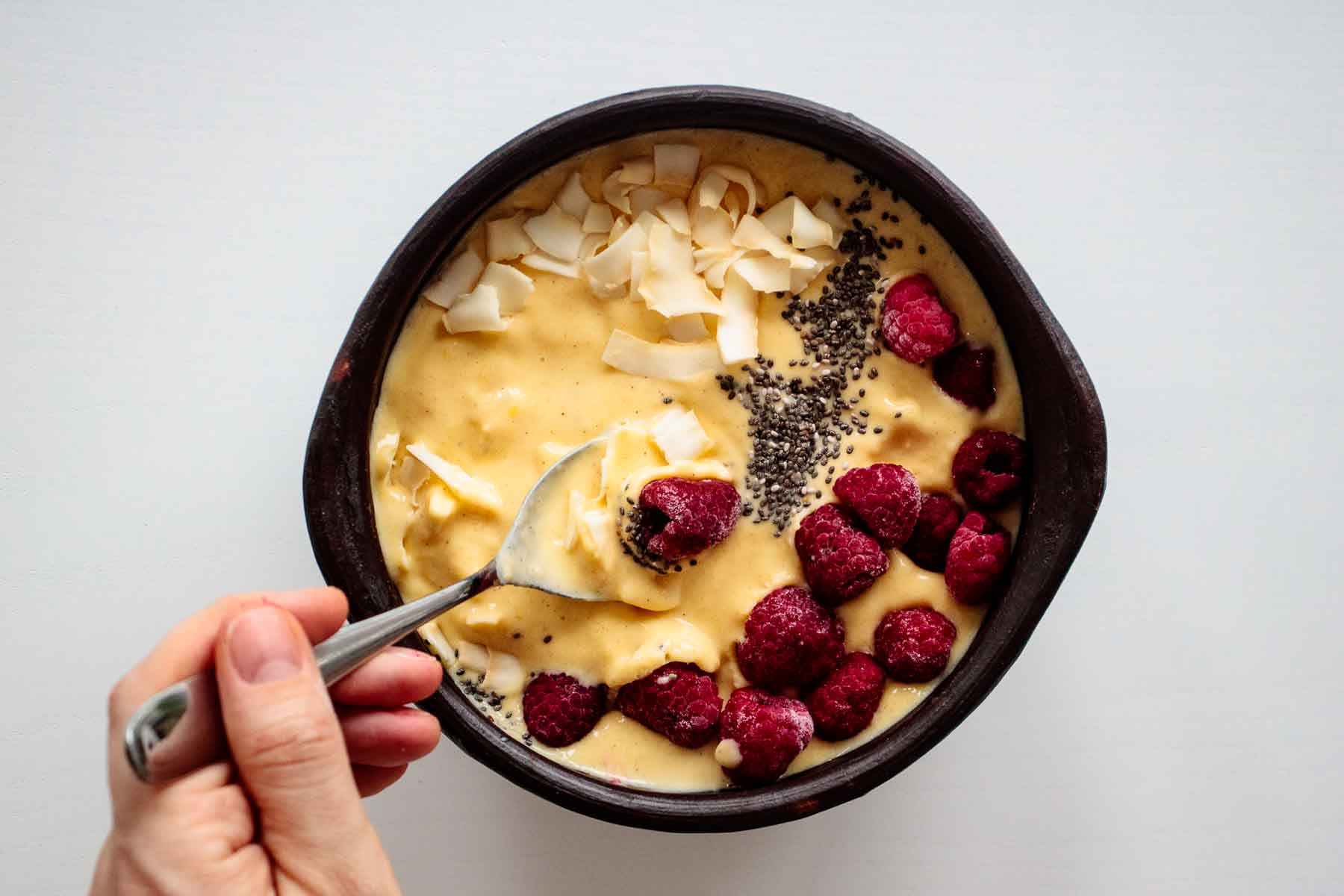 spoon scooping a breakfast bowl, with raspberry, chia and coconut flakes on top