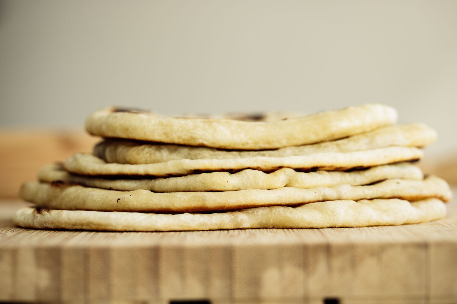 A stack of six flatbreads is placed on a wooden surface. The flatbreads appear soft and slightly golden brown, with light char marks in some areas. The background is neutral and out of focus, emphasizing the texture and layers of the flatbreads.
