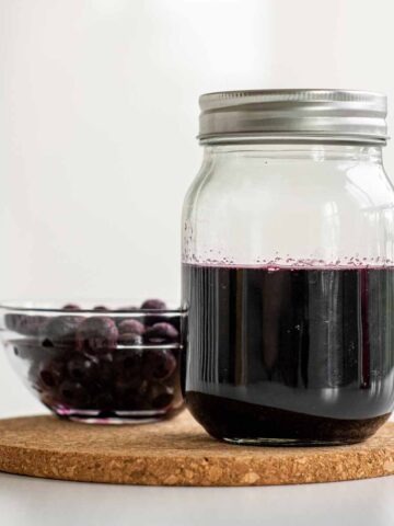 Blueberry simple syrup in a jar, with a bowl of frozen blueberries on the side.