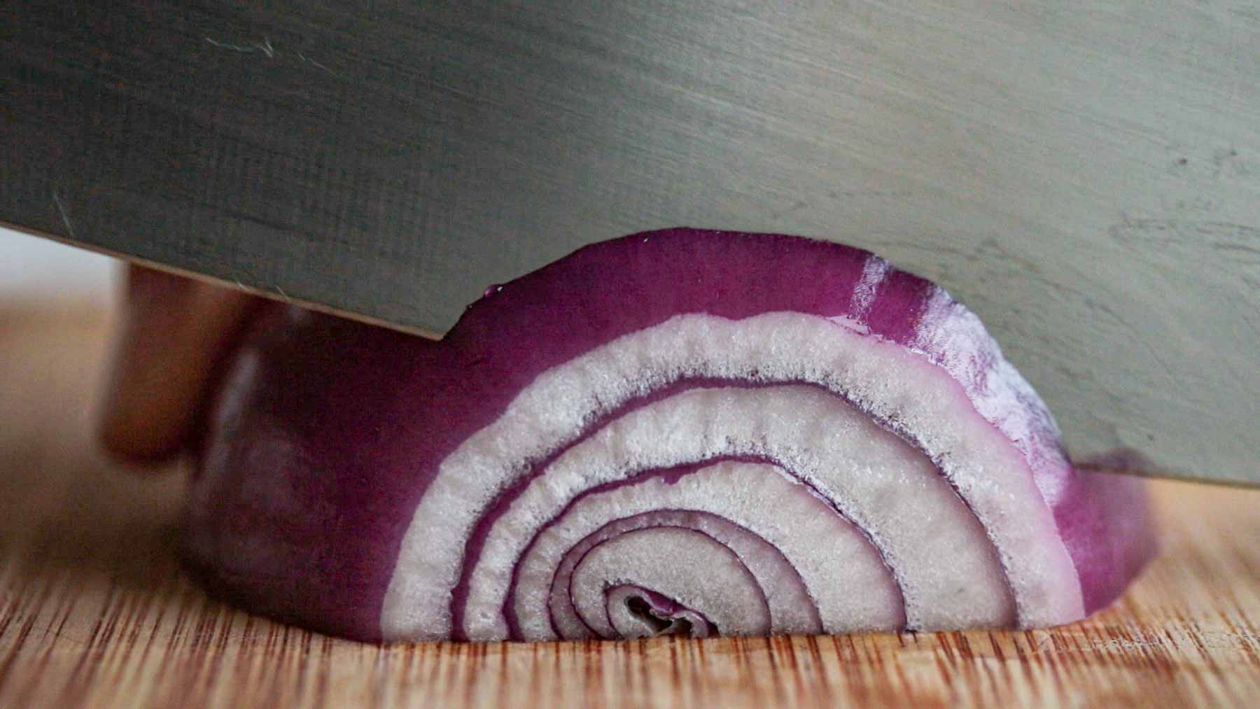 Knife slicing a red onion into half-moon slices.