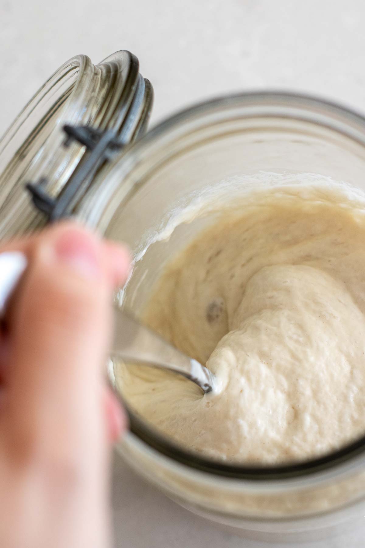 A hand mixing poolish with a spoon inside a jar.