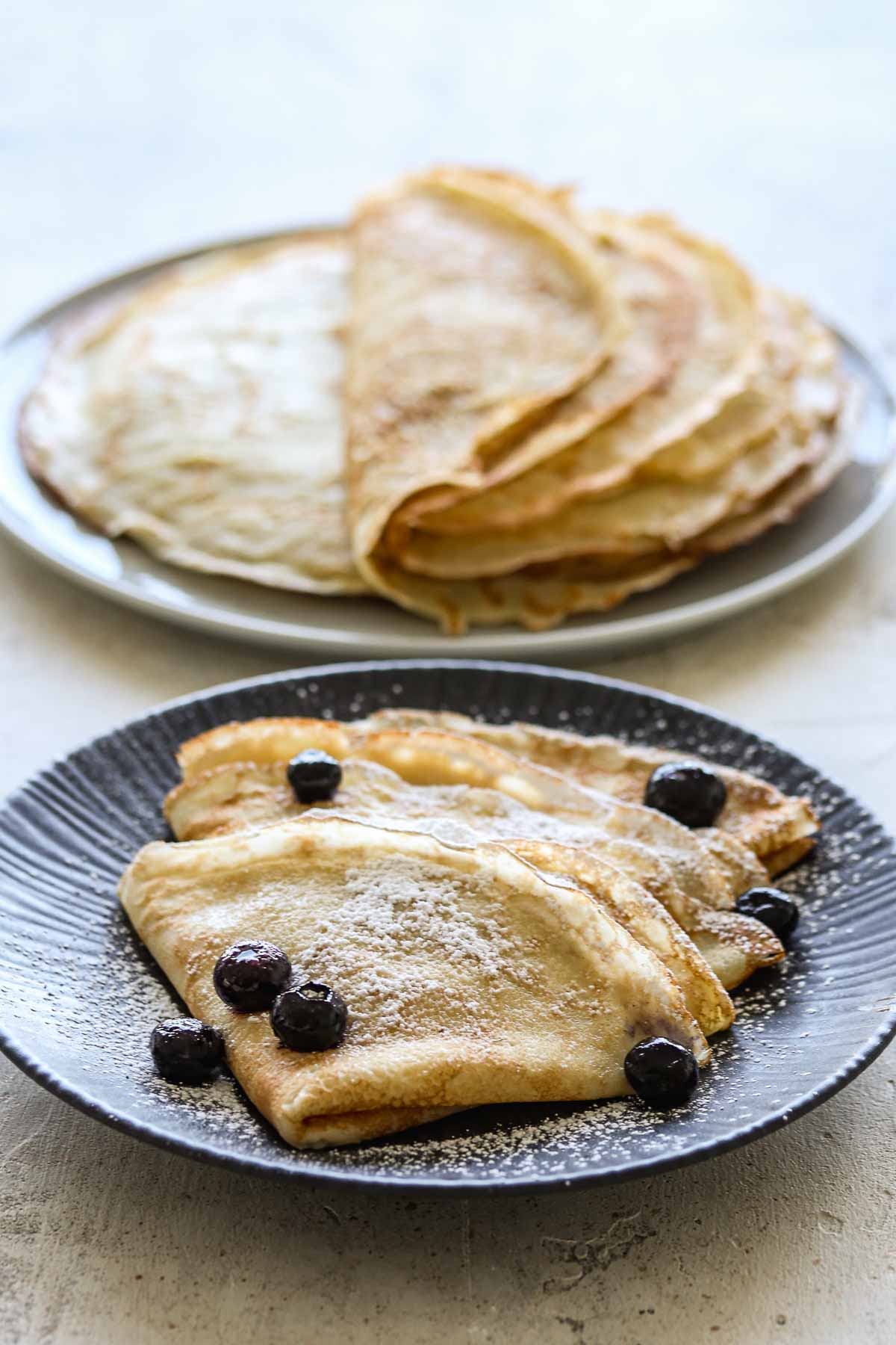 Two plates of sourdough crepes, one plain, one with icing sugar and blueberries.