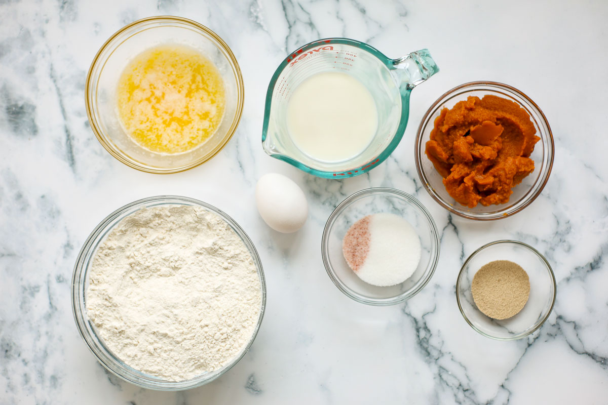 Ingredients of this recipe in glass bowls: melted butter, flour, milk, salt and sugar, yeast, pumpkin puree, and an egg.