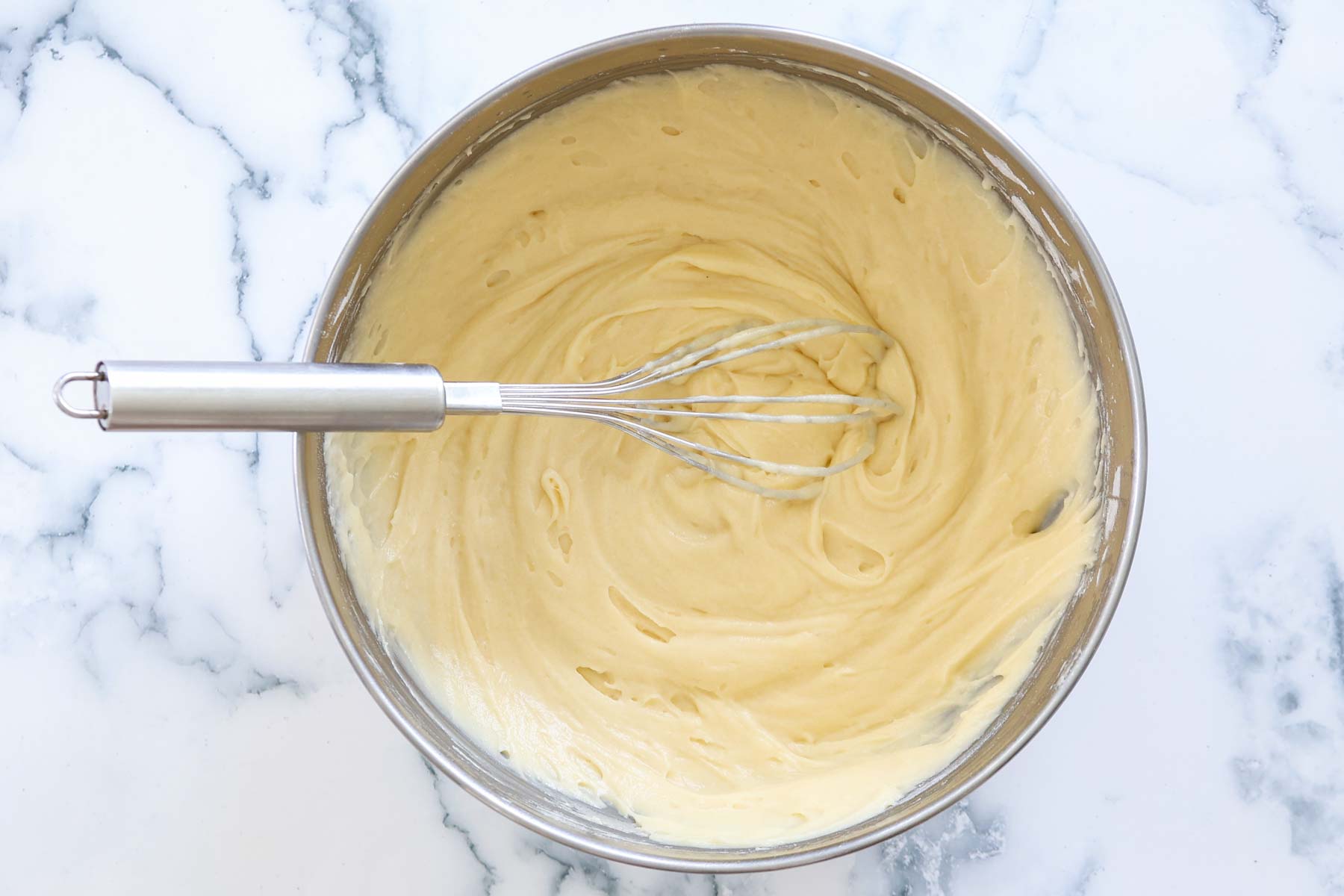 Cake batter ready with whisk, inside metal bowl.