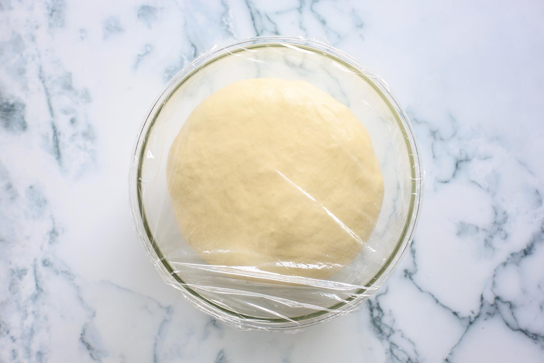 Dough after first rise, covered with plastic wrap inside glass bowl.