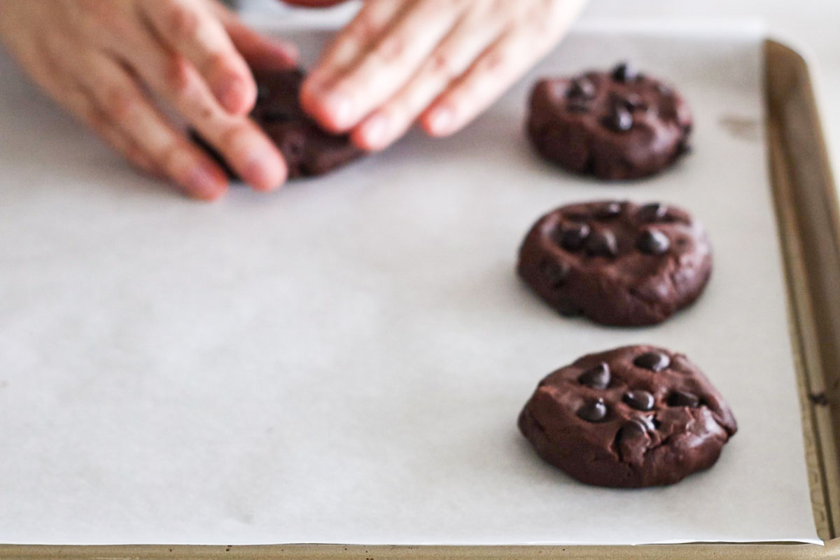 Hand flattening a chocolate dough ball on a baking sheet.