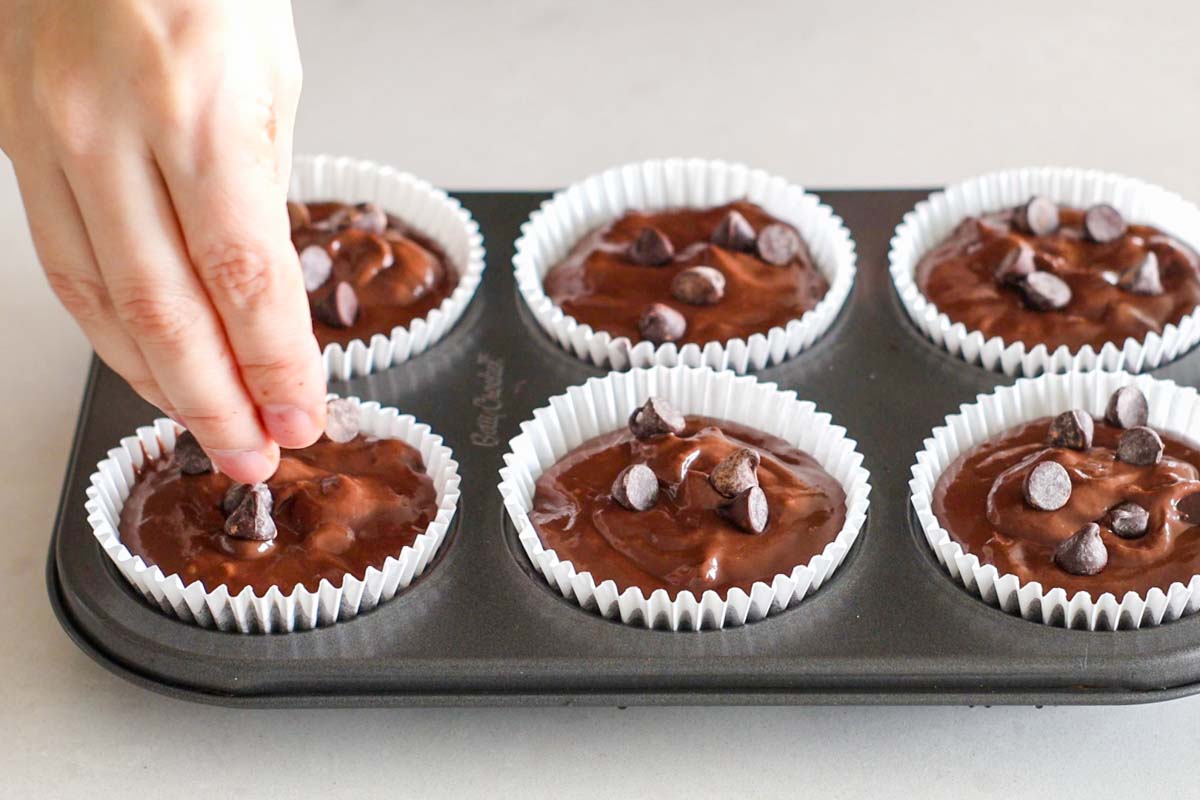 Hand adding chocolate chips on top of batter inside muffin cups.